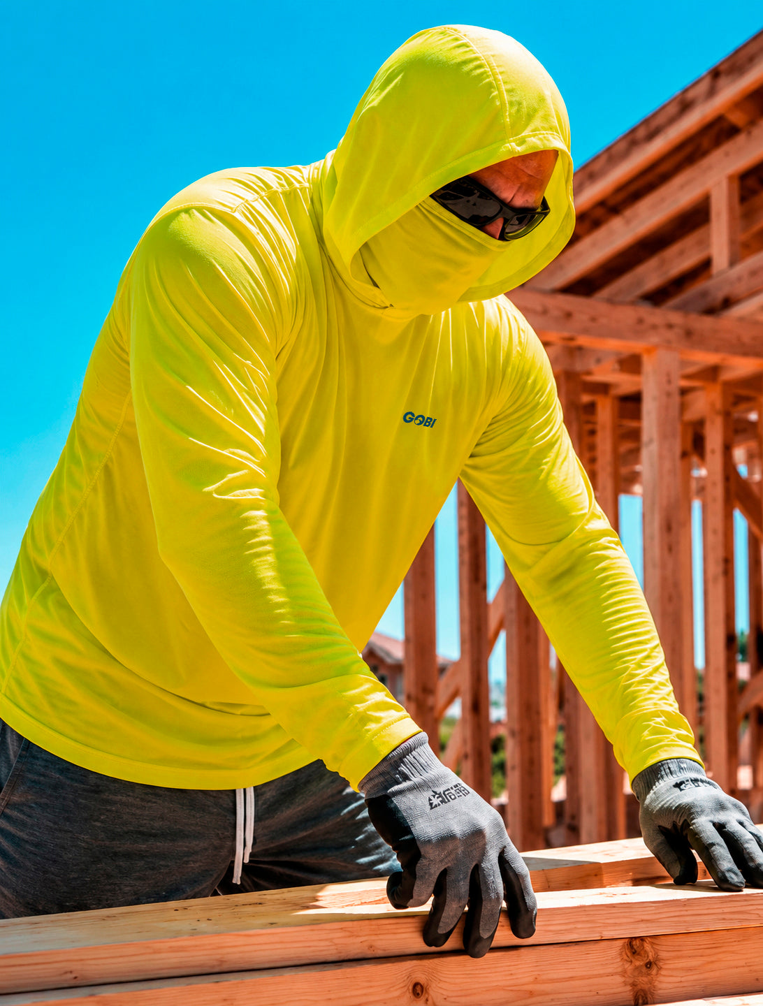 Man wearing a hi-vis yellow GOBI Helio UPF Sun Hoodie with hood and built-in face cover pulled up at a construction site, with wood framing in the background.