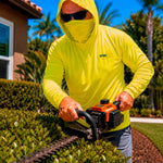 Man in a hi-vis yellow GOBI Helio UPF Sun Hoodie trimming hedges with a gas-powered hedge trimmer in a residential landscape with palm trees in the background.