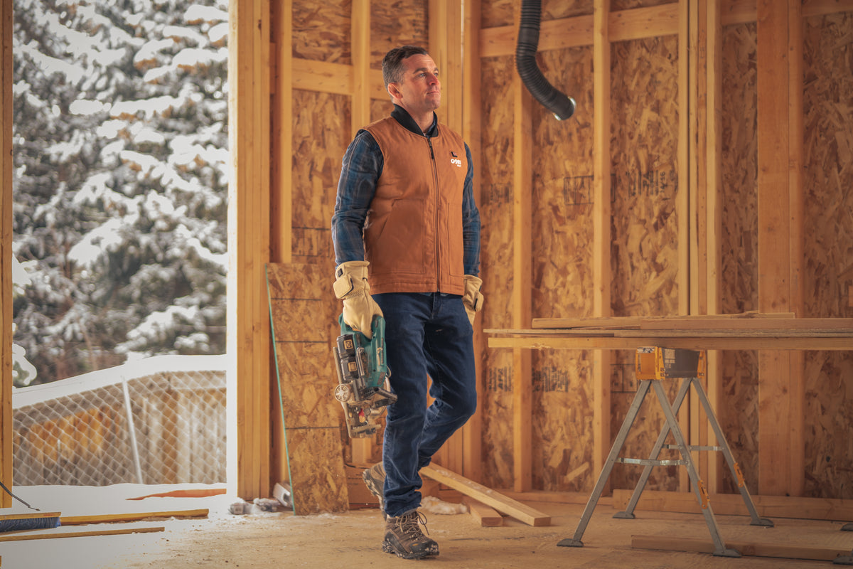Man on a winter construction site wearing the Gobi Heat Ibex heated vest in Camel over a flannel shirt, with work gloves and a tool belt, snow-covered trees visible through the open framing.