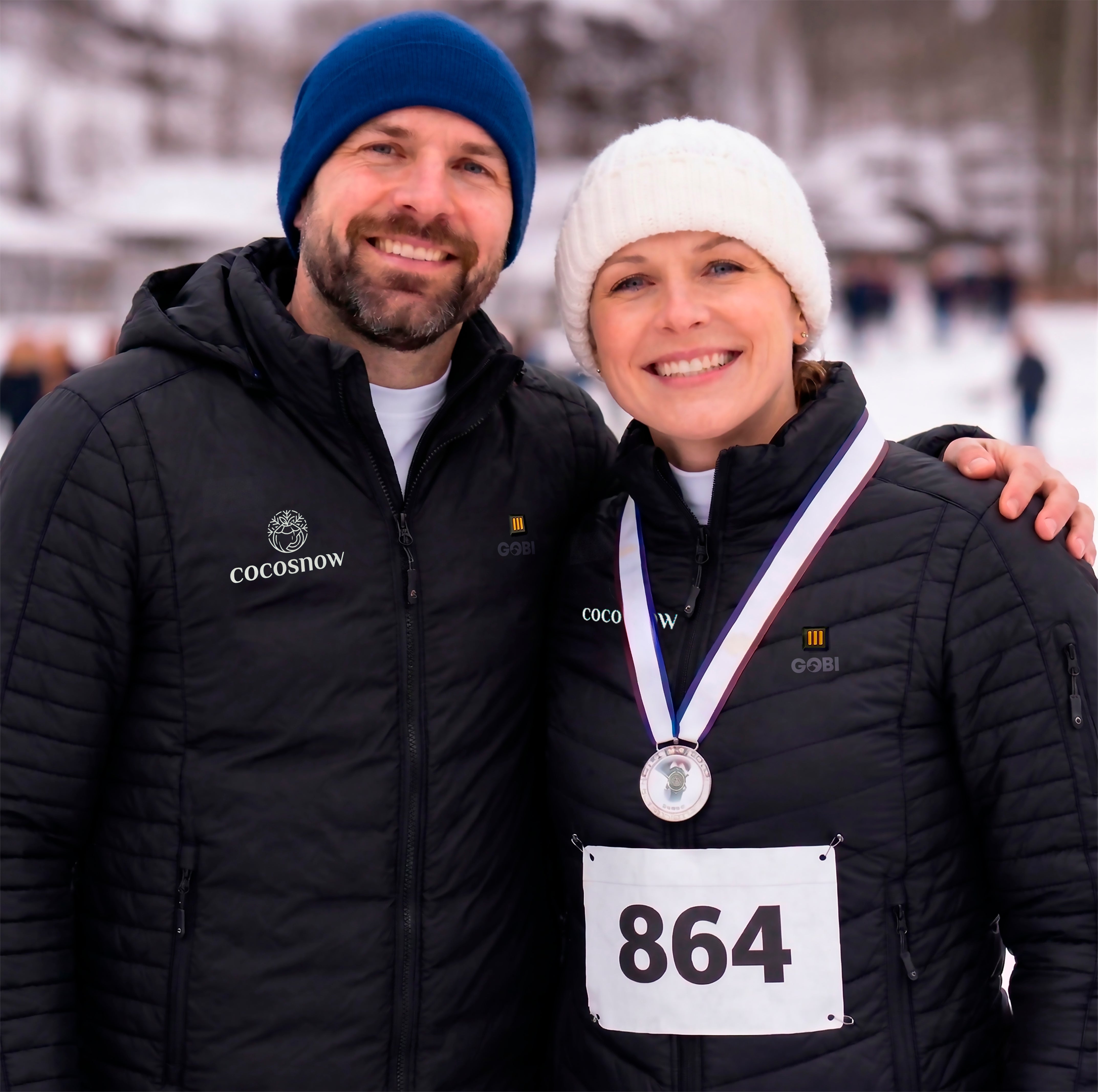 Two people in winter clothing with a medal and race number, standing outdoors in a snowy setting.