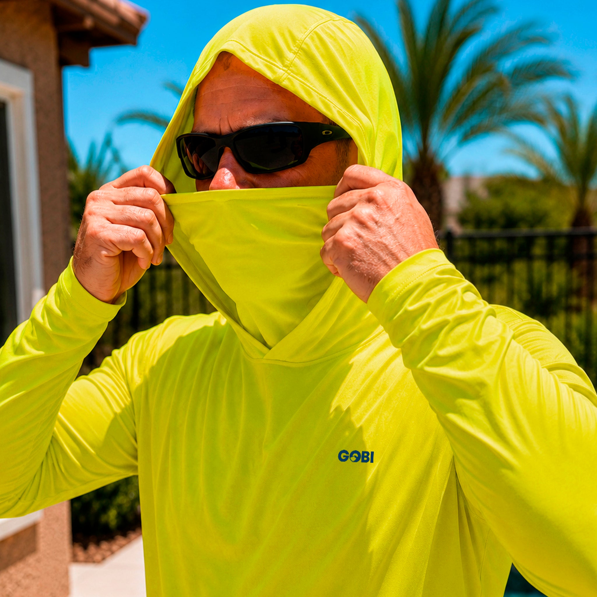 Close-up of a man pulling up the built-in face cover on a hi-vis yellow GOBI Helio UPF Sun Hoodie, wearing sunglasses outdoors with palm trees in the background.