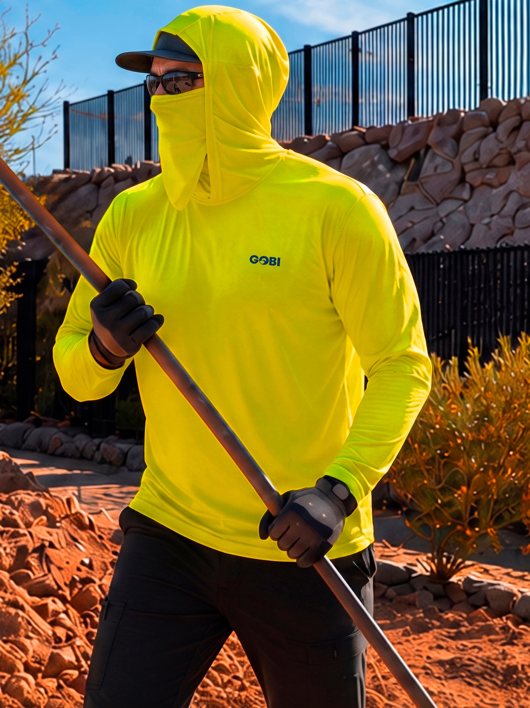 Man in a hi-vis yellow GOBI Helio UPF Sun Hoodie with hood and face cover up, holding a long-handled tool while wearing work gloves and a cap on an outdoor job site.