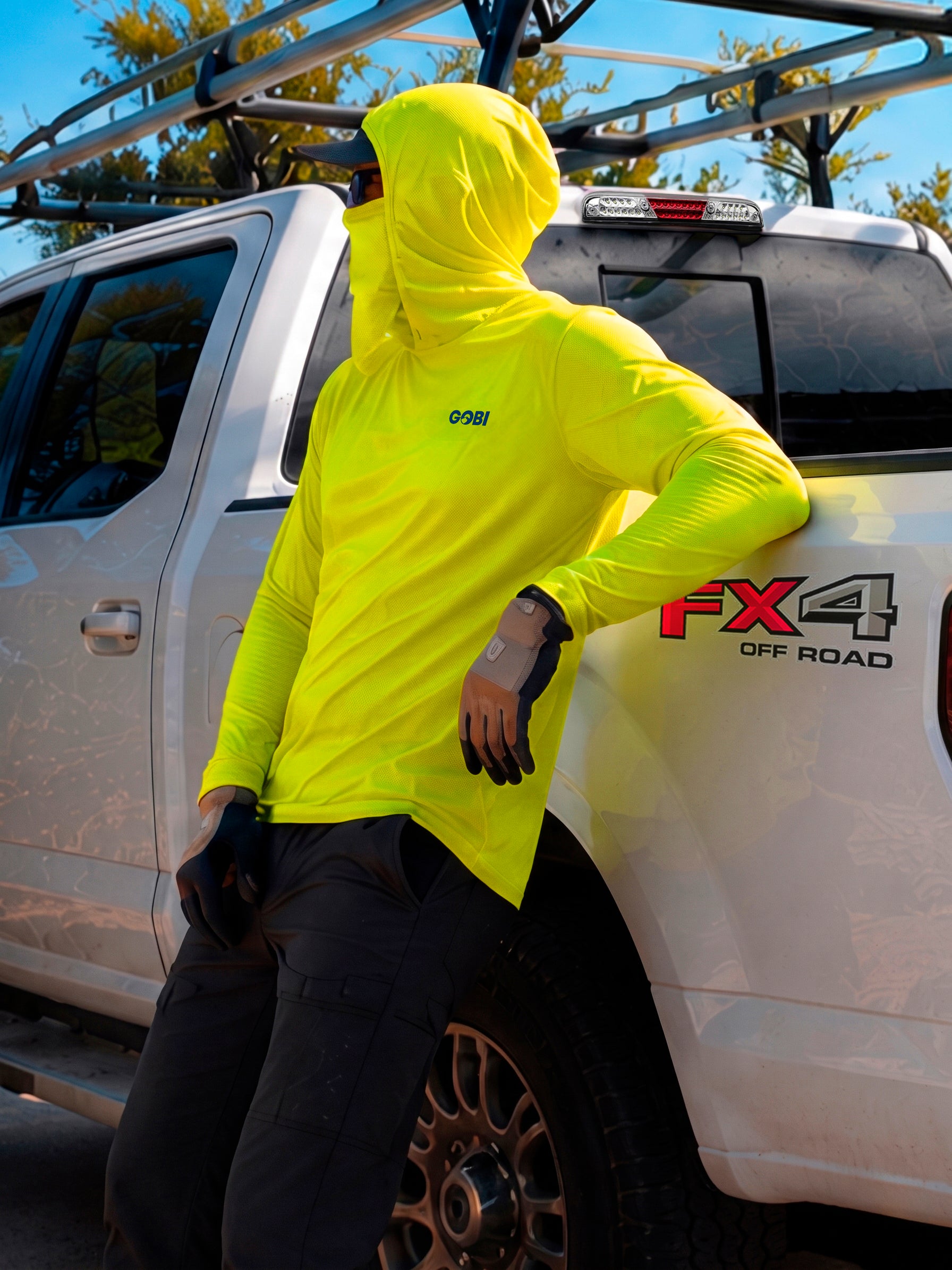 Man in a hi-vis yellow GOBI Helio UPF Sun Hoodie with hood and face cover up, leaning against a Ford FX4 Off Road pickup truck.