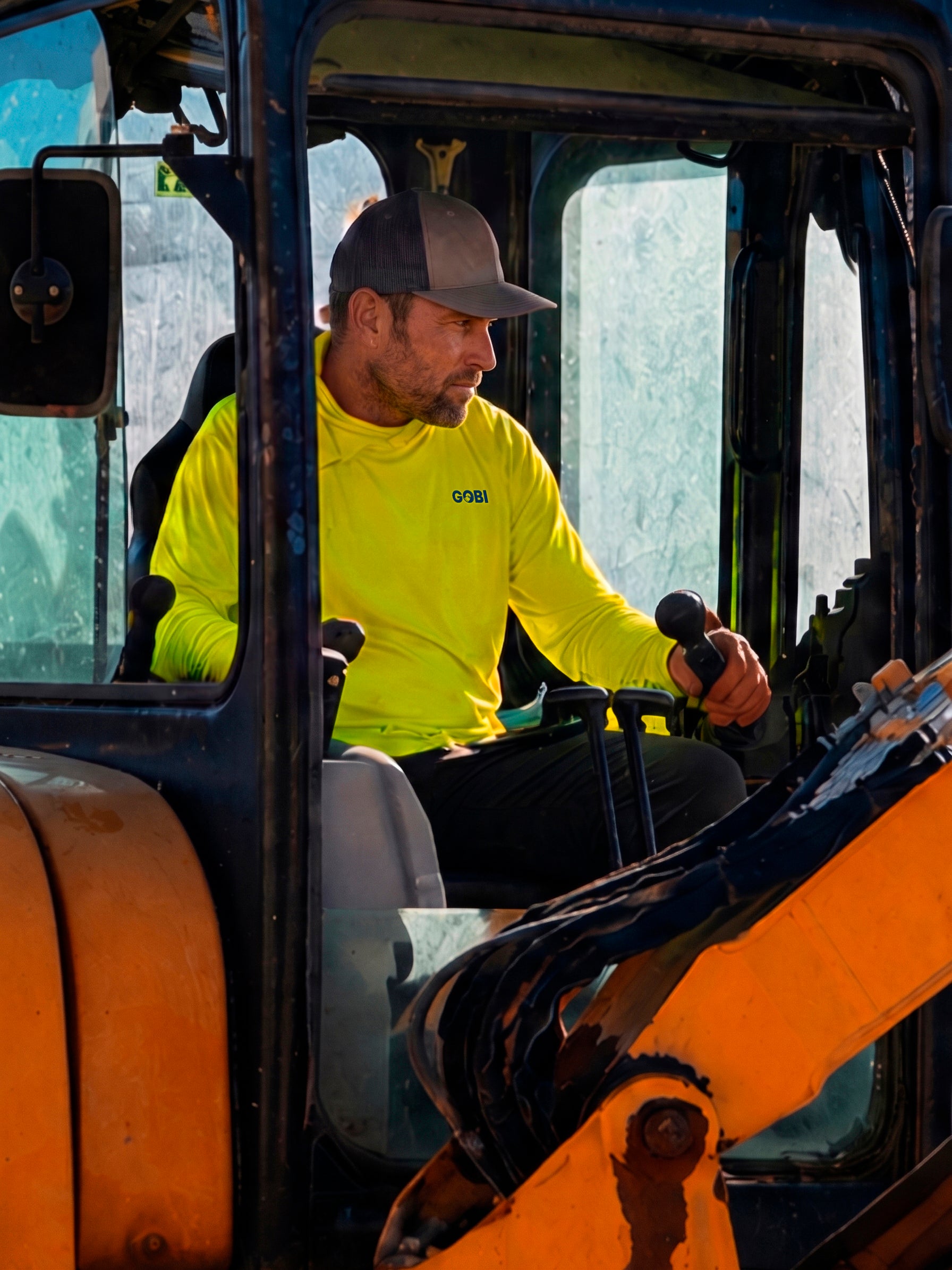 Man in a hi-vis yellow GOBI Helio UPF Sun Hoodie and trucker cap operating a forklift on a job site.